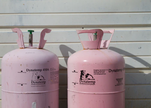Two large, pink Dynatemp gas cylinders stand side by side in front of a white metal wall. The cylinders display branding and text information in black, including details such as 'Dynatemp 410A.' Their tops are slightly open with visible valves.