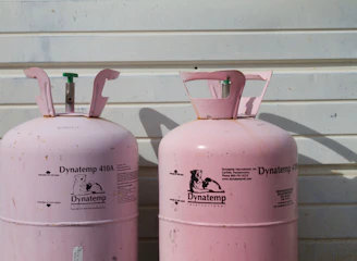 Two large, pink Dynatemp gas cylinders stand side by side in front of a white metal wall. The cylinders display branding and text information in black, including details such as 'Dynatemp 410A.' Their tops are slightly open with visible valves.