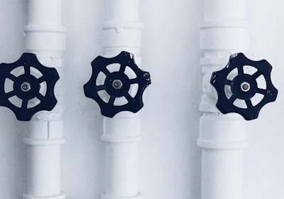Three pipes with black valve wheels are aligned vertically against a plain background. The pipes and their connecting joints are painted white, contrasting with the dark metal of the valves, which are positioned centrally on each pipe.