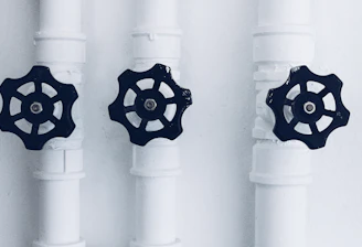 Three pipes with black valve wheels are aligned vertically against a plain background. The pipes and their connecting joints are painted white, contrasting with the dark metal of the valves, which are positioned centrally on each pipe.