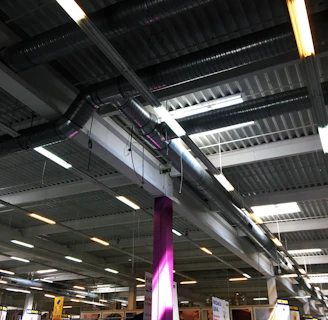 A large indoor space with an industrial-style ceiling featuring exposed ductwork and fluorescent lighting. Below, there are various goods and displays typical of a hardware or retail store with signs and product displays visible. A prominent purple column stands in the middle, surrounded by different types of merchandise.