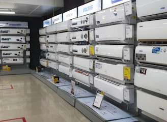 A showroom displaying a variety of air conditioners neatly arranged on two walls. Each unit is placed on shelves with price tags and brand logos visible. The floor is polished and the ceiling has fluorescent lighting.