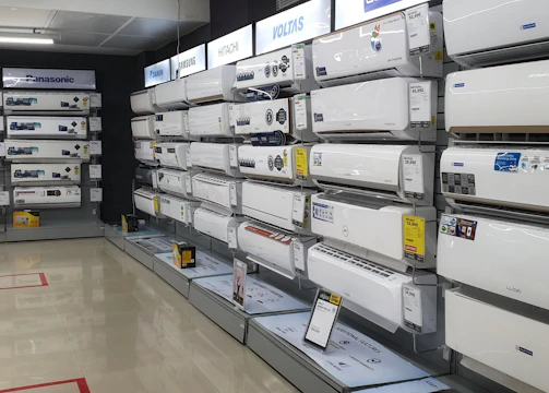 A showroom displaying a variety of air conditioners neatly arranged on two walls. Each unit is placed on shelves with price tags and brand logos visible. The floor is polished and the ceiling has fluorescent lighting.