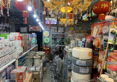 An interior of a crowded shop filled with various household and electrical items, including light bulbs, cables, and packaged goods. Red lanterns with tassels hang from the ceiling, along with ornate chandeliers. Shelves and displays are lined with boxes and products, and the space is brightly lit.