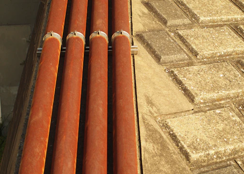 Four horizontal orange pipes are held in place by metal brackets. To the right, a surface comprising square stone tiles arranged in a geometric pattern is visible. The lighting creates warm highlights on the pipes and a textured effect on the tiles.