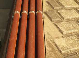 Four horizontal orange pipes are held in place by metal brackets. To the right, a surface comprising square stone tiles arranged in a geometric pattern is visible. The lighting creates warm highlights on the pipes and a textured effect on the tiles.