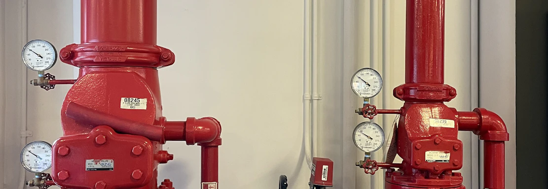Two large red industrial pipes with pressure gauges and valves are mounted on a utility wall. The pipes have labels with numbers and are connected to other smaller pipes and devices.