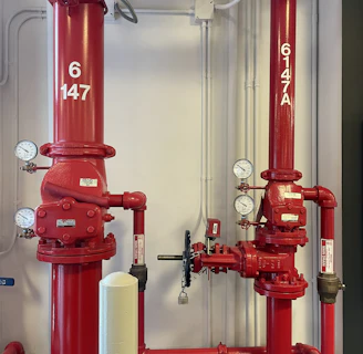 Two large red industrial pipes with pressure gauges and valves are mounted on a utility wall. The pipes have labels with numbers and are connected to other smaller pipes and devices.