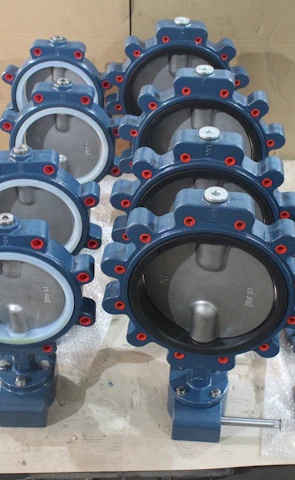 A collection of industrial butterfly valves arranged in rows on a warehouse floor. The valves are primarily metallic with blue and red accents and are placed on top of white packaging material. Additional valves and mechanical components are visible in the background alongside red fabric.