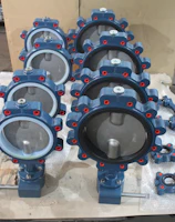 A collection of industrial butterfly valves arranged in rows on a warehouse floor. The valves are primarily metallic with blue and red accents and are placed on top of white packaging material. Additional valves and mechanical components are visible in the background alongside red fabric.