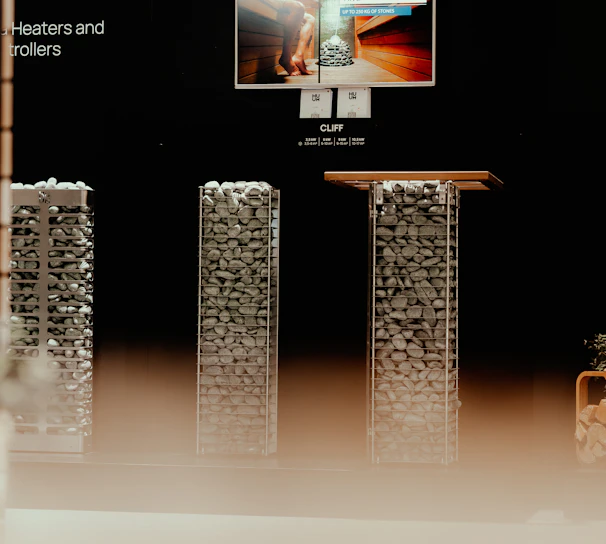 A display setup featuring a modern presentation of heating equipment. At the center, an illuminated screen shows an advertisement for sauna heaters, with a picture of a sauna interior. Below the screen, there are two tall, rectangular structures filled with evenly stacked stones, likely part of the heating equipment being showcased. The environment is dimly lit, creating a focus on the display elements.