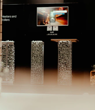 A display setup featuring a modern presentation of heating equipment. At the center, an illuminated screen shows an advertisement for sauna heaters, with a picture of a sauna interior. Below the screen, there are two tall, rectangular structures filled with evenly stacked stones, likely part of the heating equipment being showcased. The environment is dimly lit, creating a focus on the display elements.