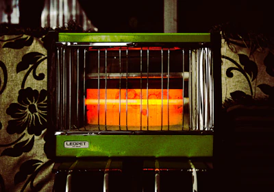 An electric heater with vertical metal bars and a glowing orange heating element is surrounded by patterned fabric upholstery. The heater has a label that reads 'LEOPET DR-622S' on a green panel.