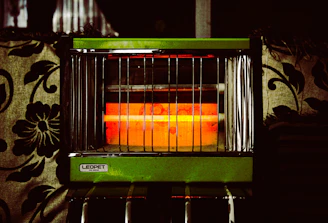 An electric heater with vertical metal bars and a glowing orange heating element is surrounded by patterned fabric upholstery. The heater has a label that reads 'LEOPET DR-622S' on a green panel.