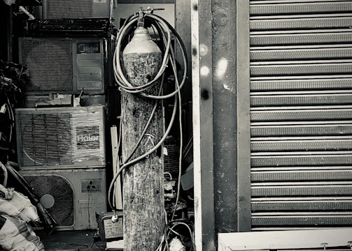 An industrial setting featuring a collection of stacked air conditioning units from various brands. A tall cylinder with hoses wrapped around it stands prominently in the foreground. The setting appears to be a cluttered storage or workshop space, with a partially visible metal roller shutter door on one side.