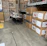 A warehouse setting with stacks of items in cardboard boxes. Some boxes contain outdoor furniture like chairs. There are also large industrial fans or air conditioning units wrapped in plastic on the left side. The floor is concrete, and there's a marked yellow area.