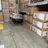 A warehouse setting with stacks of items in cardboard boxes. Some boxes contain outdoor furniture like chairs. There are also large industrial fans or air conditioning units wrapped in plastic on the left side. The floor is concrete, and there's a marked yellow area.