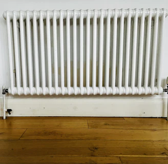 A white, wall-mounted radiator with vertical bars is fixed to a plain white wall above a light wooden floor. The baseboard and pipe fittings are also visible, with some signs of wear and discoloration on the radiator.