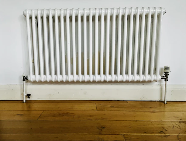 A white, wall-mounted radiator with vertical bars is fixed to a plain white wall above a light wooden floor. The baseboard and pipe fittings are also visible, with some signs of wear and discoloration on the radiator.