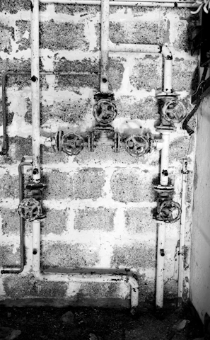A black and white image showing a system of old, rusty pipes and valves mounted on a rough brick wall. The scene is industrial and appears to be part of a plumbing system. The pipes are arranged horizontally and vertically with several turning wheels for controlling the flow.