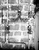 A black and white image showing a system of old, rusty pipes and valves mounted on a rough brick wall. The scene is industrial and appears to be part of a plumbing system. The pipes are arranged horizontally and vertically with several turning wheels for controlling the flow.