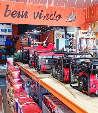 A hardware or general store interior featuring several generators displayed prominently on a central counter. Shelves are stocked with various products including paint cans, tools, and other equipment. Straw hats are hung decoratively above the counter. The store is well-lit and organized, with an inviting atmosphere suggested by a 'Seja bem-vindo' sign, which means 'welcome' in Portuguese.