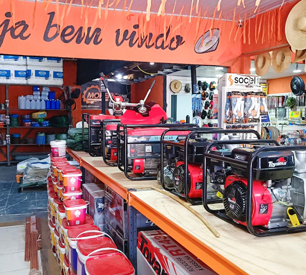 A hardware or general store interior featuring several generators displayed prominently on a central counter. Shelves are stocked with various products including paint cans, tools, and other equipment. Straw hats are hung decoratively above the counter. The store is well-lit and organized, with an inviting atmosphere suggested by a 'Seja bem-vindo' sign, which means 'welcome' in Portuguese.