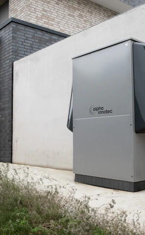 A modern, rectangular air conditioning or heating unit with the logo 'alpha innotec' is installed outside a building with a dark gray brick exterior. The unit is positioned against a light concrete wall, and there is overgrown grass along the edge of the pavement.