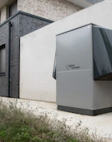 A modern, rectangular air conditioning or heating unit with the logo 'alpha innotec' is installed outside a building with a dark gray brick exterior. The unit is positioned against a light concrete wall, and there is overgrown grass along the edge of the pavement.